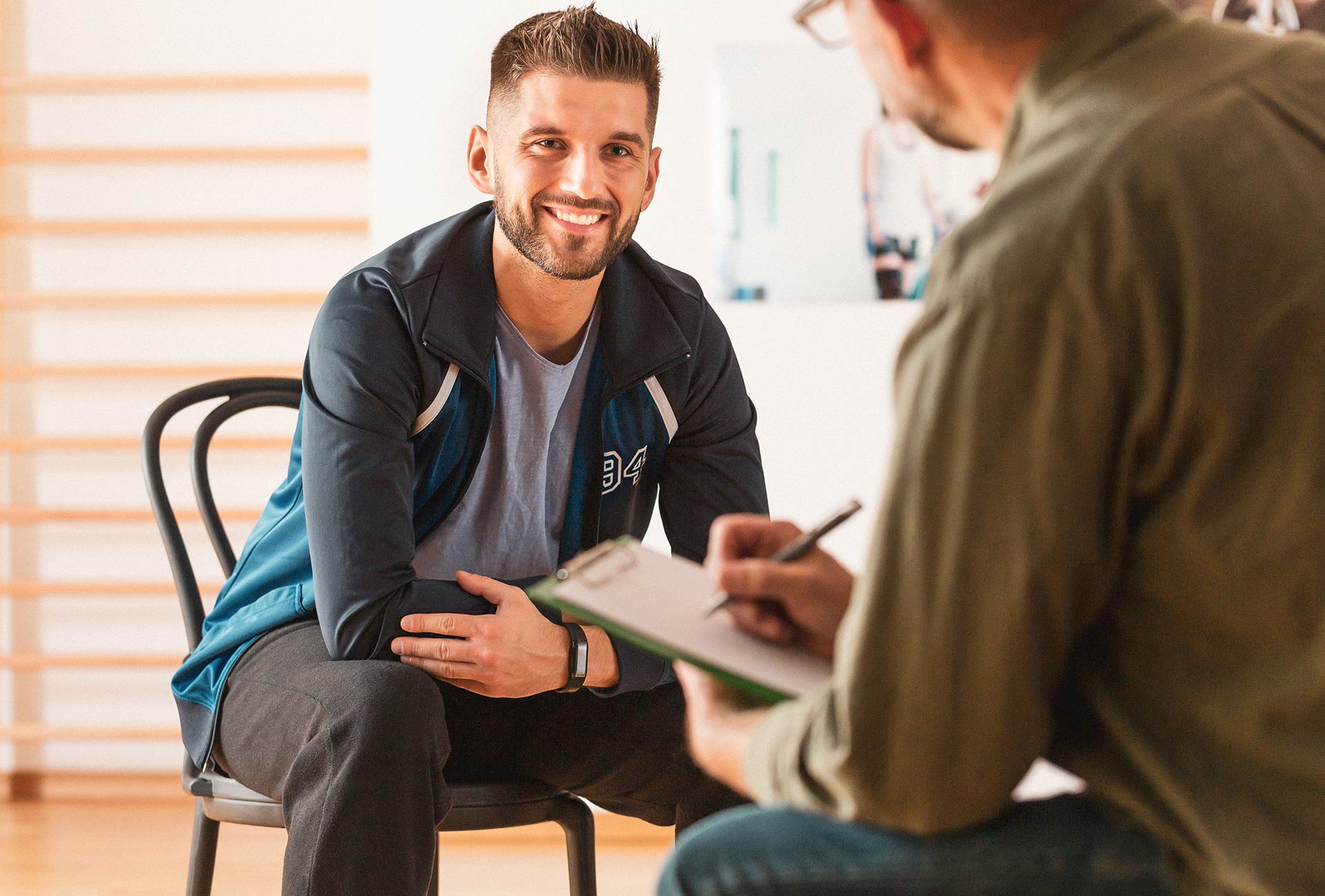 A smiling man sits on a chair wearing a casual jacket, engaging in a one-on-one counseling session. In the foreground, a therapist holds a clipboard and takes notes, symbolizing the supportive environment of outpatient therapy.