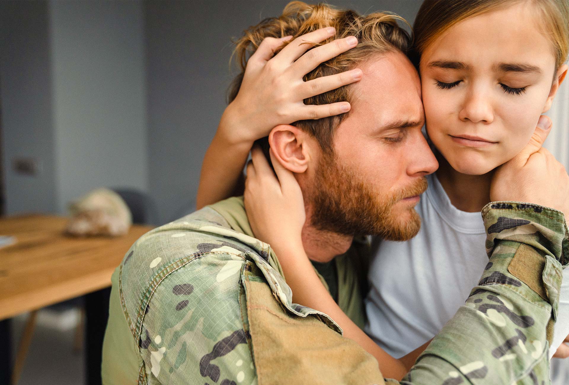 A soldier in a camouflage uniform embraces a young girl with a heartfelt expression of comfort and connection. The soldier has his eyes closed, conveying deep emotion, while the girl rests her hands on his head, signifying mutual support and understanding. The image highlights themes of trauma, healing, and emotional resilience.